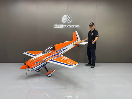 Person standing next to an orange and white RC airplane in a showroom with a brand logo on the wall.
