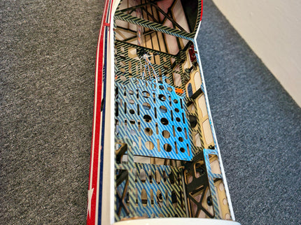 A close-up view of a model airplane fuselage showing the open cockpit interior with a blue wing, carbon fiber bracing, and red and white body panels.