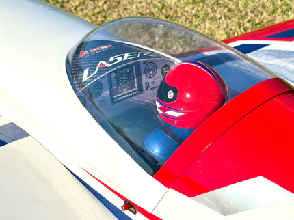 Close-up of a red and white RC airplane cockpit with a blue and red helmet visible inside under a clear canopy. The canopy has 'LASER' branding printed on it.