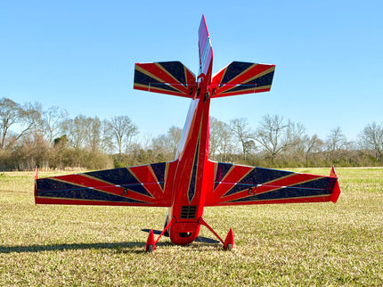 A red, white, and blue high-wing airplane with a Union Jack flag pattern on the wings and tail, parked on a grassy field under a clear blue sky.