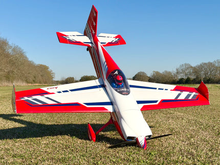 A red, white, and blue RC model high-performance racing airplane parked on a grassy field under a clear blue sky.