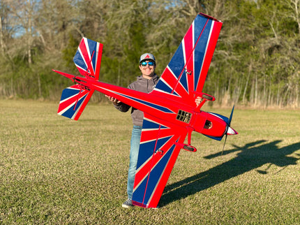 A person standing outdoors in a grassy field holds a large red, white, and blue model airplane with a Union Jack flag design on the wings and tail. Trees are visible in the background, and the person is wearing a cap, sunglasses, gray jacket, and jeans.