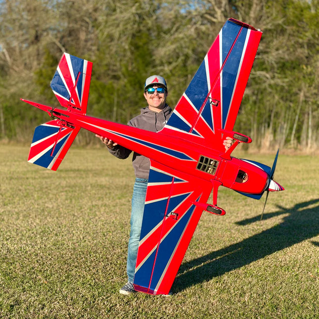 A person standing outdoors in a grassy field holds a large red, white, and blue model airplane with a Union Jack flag design on the wings and tail. Trees are visible in the background, and the person is wearing a cap, sunglasses, gray jacket, and jeans.