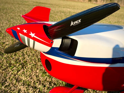 Close-up of a red, white, and blue model airplane with black propellers, photographed outdoors on grass. The fuselage has white stars and stripes, and the propellers show the Apex Wings logo.
