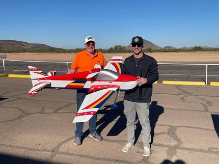 Two men holding a large model airplane on a tarmac with mountains in the background