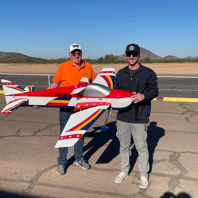 Two men holding a large model airplane on a tarmac with mountains in the background