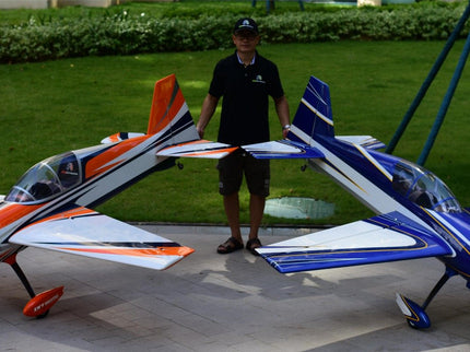 Man standing between two large model airplanes on a concrete surface with grass and trees in the background.