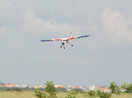Phoenix Model Domino 62” Trainer flying in a clear sky with trees and buildings in the background