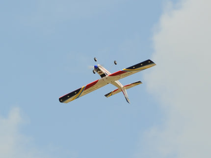Phoenix Model Domino 62” Trainer flying against a blue sky with some clouds.