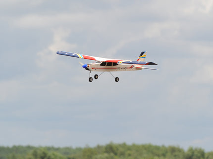 Phoenix Model Domino 62” Trainer Model airplane flying in the sky with trees in the background