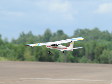 Phoenix Model Domino 62” Trainer flying over a runway with trees in the background