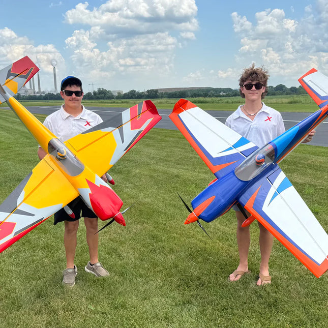 Two people holding large model airplanes on a grassy field with a clear sky.