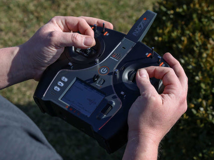 A close-up of a person's hands holding a black and gray radio transmitter controller outdoors with blurred green foliage in the background.