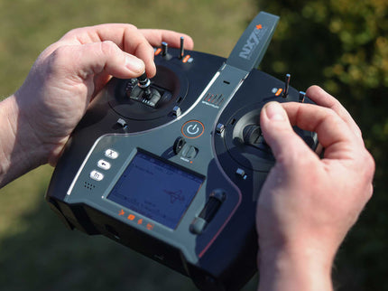 A close-up of a person's hands holding a black and gray RC airplane transmitter controller outdoors with green grass in the background. The controller has dual joysticks, a small screen, and orange button accents.