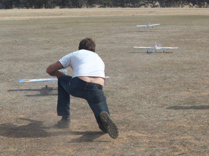 Person launching a Phoenix Model PH006-SCANNER 57" model airplane on a grassy field with another plane in the background.