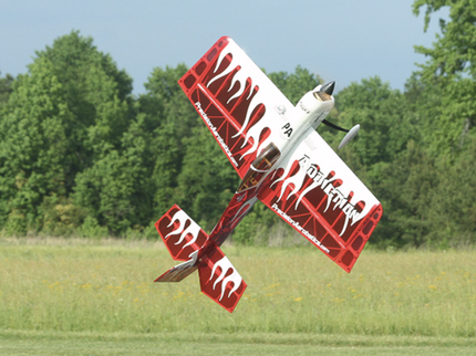 Red and white model airplane flying over a grassy field with trees in the background