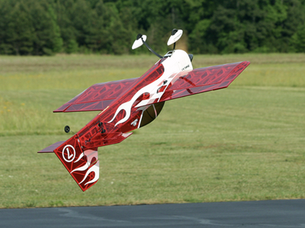 Red and white model airplane flying over a grassy field with trees in the background