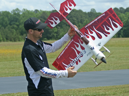 Man holding a red and white model airplane with flames on a grassy field.