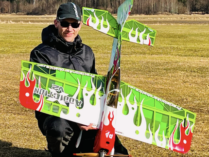Person holding a green and red model airplane in an open field with trees in the background