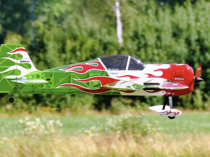 Model airplane with green and red design flying over a grassy field with trees in the background
