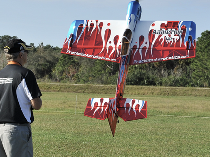 Man controlling a red and white model airplane in an open field with trees in the background