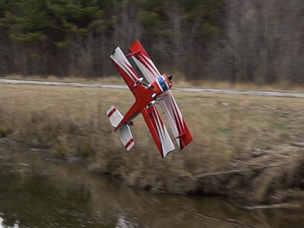 Red and white model airplane flying over a body of water with trees in the background