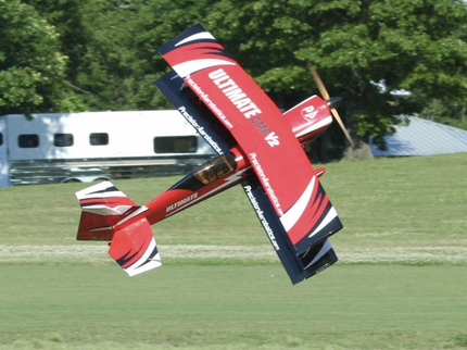 Red and black stunt airplane performing a maneuver in a grassy field with trees in the background.