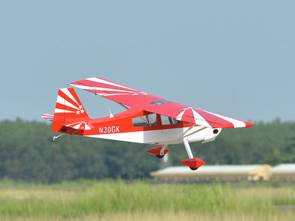 Red and white Phoenix Model DECATHLON airplane flying over a grassy field with trees in the background