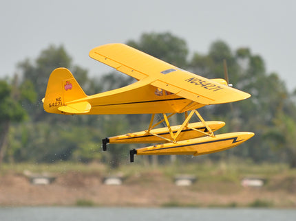 Yellow Phoenix Model PIPER J3 CUB  seaplane flying over a body of water with trees in the background