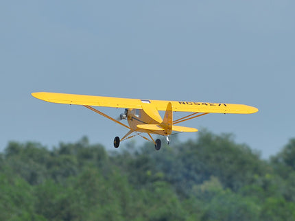 Yellow Phoenix Model PIPER J3 CUB  flying against a clear blue sky with green trees below.