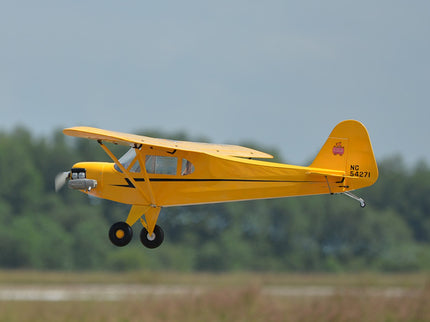 Yellow Phoenix Model PIPER J3 CUB airplane flying over a grassy field with trees in the background