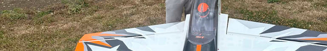 Probuild Team Pilot Ashton Boycott in a red apex shirt and black cap stands behind two white, black, and orange remote control model airplanes in an outdoor grassy field.