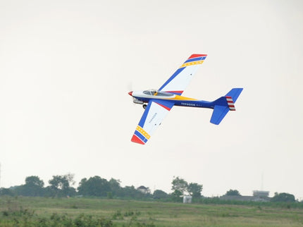 Colorful Phoenix Model TYPHOON 59" Size ARF airplane flying over a grassy field with trees in the background